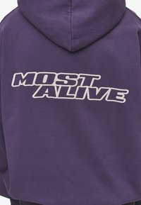 Sudadera púrpura que presenta un gran texto impreso en blanco en la parte posterior que dice "MOST ALIVE". La tela parece suave con un ajuste casual y capucha.