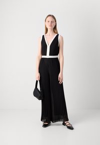 Jumpsuit noir plissé avec un décolleté en V profond, accent blanc à la taille, associé à un sac à main noir et des chaussures noires à bout carré.