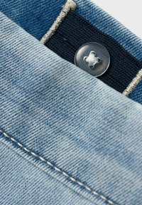 Svetlomodrá denim látka s viditeľným strieborným gombíkom, kontrastným tmavým opaskom a bielym prešitím pozdĺž okrajov.