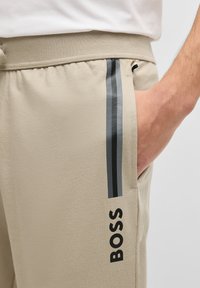 Beige joggers met een zachte, gestructureerde afwerking hebben zwarte en grijze zijstrepen. Het merkembleem is in vet zwart op de bovenbeen weergegeven.