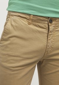 Khaki chinos vyrobené z bavlny s hladkým povrchem. Mají knoflíkový zapínání, poutka na opasek a boční kapsy s čistým designem.