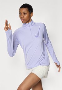 Vaaleanlila pitkähihainen urheilu paita, jossa on neljännesvetoketju ja peukaloaukot. Rinnassa on valkoinen Nike-logon swoosh.
