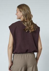 Bordeaux ærmeløs top med løs pasform, der har en høj hals og blød tekstur. Matcher med beige bukser med samlet talje.