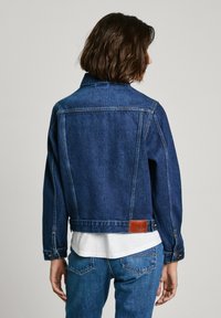 Giacca di jeans blu scuro con taglio classico, caratterizzata da un'etichetta in pelle sul retro, polsini con bottoni e orlo dritto.