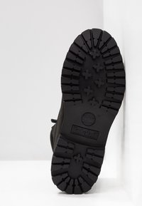 Semelle d'une botte Timberland noire montrant un motif de bande profonde et le logo Timberland embossé ainsi que le symbole de l'arbre sur un fond blanc.