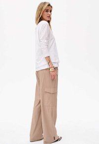 Weißes Langarmoberteil kombiniert mit beigen, weiten Cargo-Hosen mit Seitentaschen und lockerer Passform, stylisch ergänzt durch goldene Armbänder und gemusterte Schuhe.