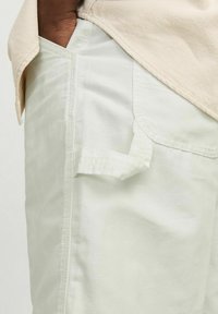 Pantaloni bianchi in cotone con una tasca laterale e un piccolo anello in tessuto. Il tessuto ha una texture liscia con leggere pieghe.