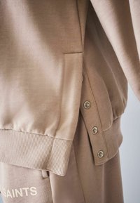 Beige-collegepaita, valmistettu pehmeästä kankaasta, jossa on ribbineuloshelma, sivutasku ja sivussa painonappiyksityiskohdat. Hienovaraiset tekstuurikontrastit.