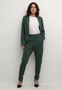 Tmavě zelený přizpůsobený blazer a odpovídající kalhoty v kombinaci s bílým tričkem. Vyznačuje se elegantními liniemi a jemnou, hladkou texturou. Černé podpatky.