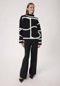 Suéter negro de gran tamaño con líneas abstractas blancas, cuello alto y textura suave. Combinado con pantalones negros a medida y zapatos negros de punta.