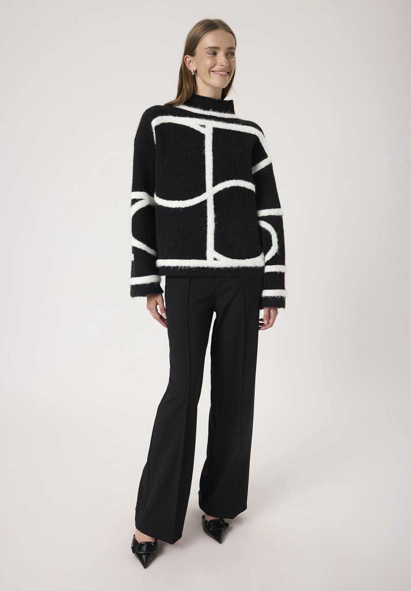 Suéter negro de gran tamaño con líneas abstractas blancas, cuello alto y textura suave. Combinado con pantalones negros a medida y zapatos negros de punta.