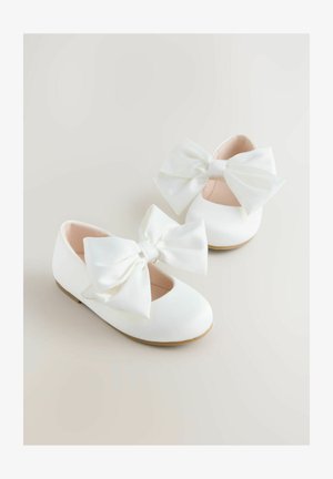 Baltas baleta dzīvokļkurpes ar lielām satīna lokām priekšpusē, apaļu purngalu un mīkstu iekšējo oderējumu; minimālistisks dizains ar gludu tekstūru.