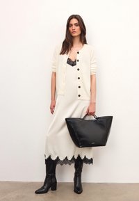 White dress with lace hem, black cardigan, and knee-high black leather boots. Holding a large black leather tote with a smooth finish.
