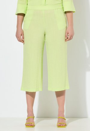 Limoengroene, gestructureerde, wijde cropped broek met zijzakken, gecombineerd met gele sandaaltjes met bandjes, gedragen door een staand persoon.