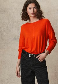 Helles oranges Strickpullover mit langen Ärmeln, kombiniert mit schwarzen Jeans im Zebra-Muster und einem schwarzen Gürtel mit einer silbernen Schnalle.