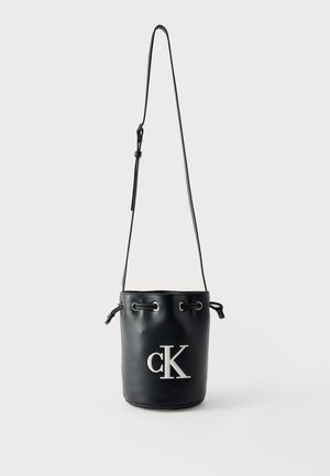 Sac seau en cuir noir avec bandoulière réglable et logo blanc "cK" sur le devant, fermeture par cordon en haut.