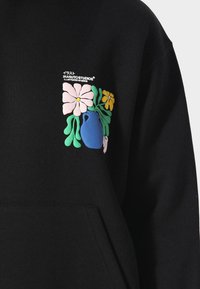 Mustat hupparit, joissa on värikäs graafinen kuvasto, jossa on vaaleanpunaisia ja keltaisia kukkia sekä sininen maljakko vasemmalla rintapielessä. Pehmeää kangasta, jossa on kenguru-tasku.