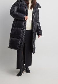 Schwarze Steppjacke mit glänzender Oberfläche, Reißverschluss und Druckknopfverschluss, großer Kapuze, langem Schnitt und seitlichen Schlitzen; getragen über einem cremefarbenen Pullover und schwarzen Jeans.