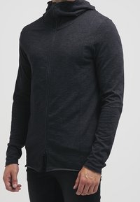 Zwarte hoodie met ritssluiting, gemaakt van zacht, gestructureerd materiaal. Heeft een hoge kraag, lange mouwen en een getailleerd ontwerp met subtiele naden.