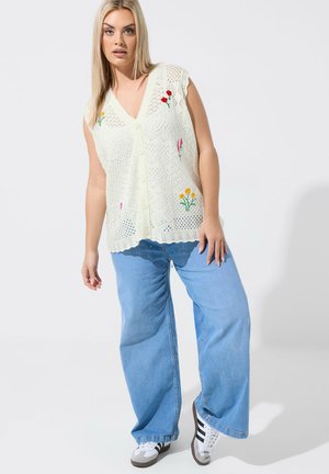 Blonde Frau trägt eine ärmellose weiße Strickweste mit Blumenstickerei, hellblaue Jeans mit weitem Bein und weiße Turnschuhe mit schwarzen Streifen.