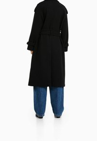 Czarny płaszcz trench z wełny z paskiem w talii, długimi rękawami i klasycznym kołnierzem, zestawiony z luźnymi niebieskimi dżinsami.
