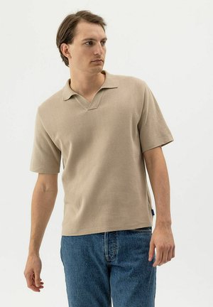 Fiatal férfi bézs rövid ujjú kötött galléros pólóban és kék farmert viselve, balra néz egy egyszínű világos háttér előtt.