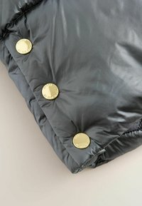 Chaqueta acolchada gris en la esquina con botones a presión dorados. El material parece suave y reflectante, con secciones acolchadas que tienen un ligero brillo.