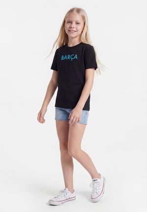 Uśmiechnięta dziewczyna z długimi blond włosami, ubrana w czarną koszulkę "BARÇA", jasne dżinsowe szorty oraz białe trampki, idąca na białym tle.
