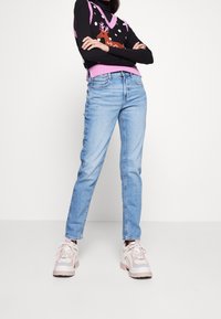 Helle blaue, hoch taillierte Jeans aus Denim, kombiniert mit einem schwarzen Strickwesten mit einem Rentierdesign und pinken Akzenten, getragen mit klobigen Sneakers.