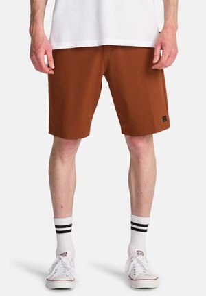 Bruine shorts met een rechte snit, gemaakt van lichtgewicht stof. Draagt witte schoenen en gestreepte sokken, staand tegen een effen achtergrond.