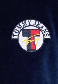 Patch con il logo Tommy Jeans caratterizzato da un design rosso e bianco su uno sfondo blu scuro, con testo ricamato e una linea di accento gialla.