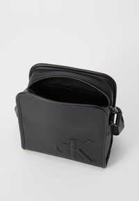 Black leather crossbody bag with a zippered top, embossed logo on the front, and a slim, structured shape. Interior features a single pocket.