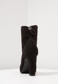 Černé velurové kotníkové boty na vysokém podpatku s zipem vzadu a hladkým povrchem, umístěné na jednobarevném pozadí.