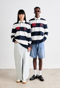 Stripete rugby-stil skjorter i marineblå og hvit med røde detaljer. Kvinnen har på seg en lang lys denimskjørt; mannen har på seg denimshorts og svarte sko.