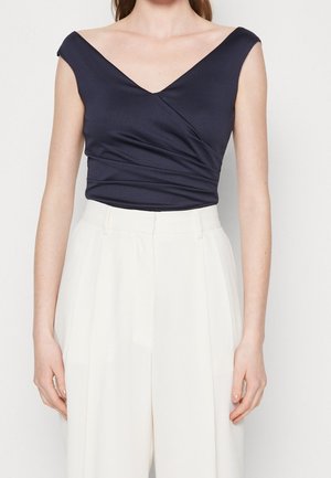 Femme portant un haut bleu marine sans manches style cache-cœur avec un profond décolleté en V et un pantalon blanc large taille haute.