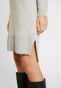 Abito grigio lavorato a maglia con orlo a coste, spacchi laterali e discreto dettaglio del logo. Il tessuto sembra morbido, con una trama liscia. Stivali neri al ginocchio.