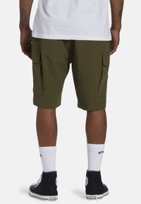 Shorts cargo vert olive avec poches latérales, fabriqués dans un tissu léger, associés à des baskets montantes noires et des chaussettes blanches.