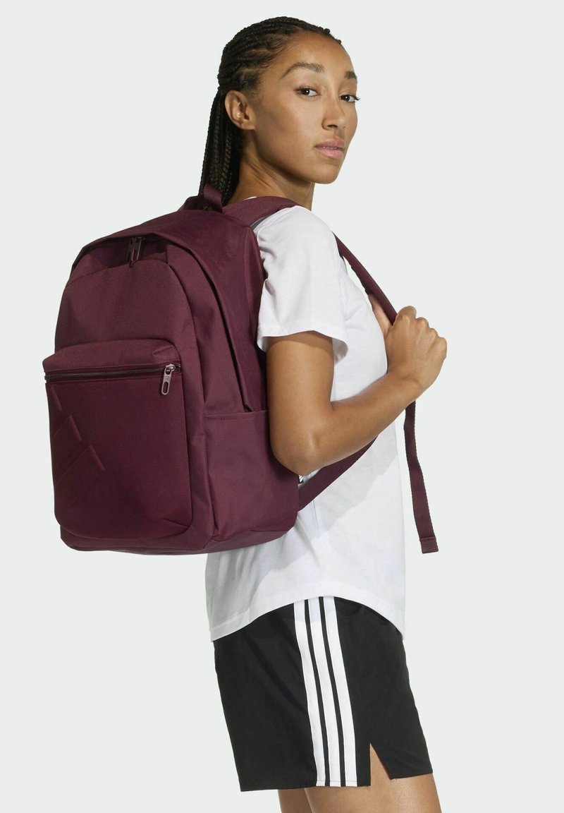 Mochila de tela color burdeos con un bolsillo frontal con cremallera, correas ajustables y un detalle de patrón geométrico. Modelo con camiseta blanca y pantalones cortos negros.