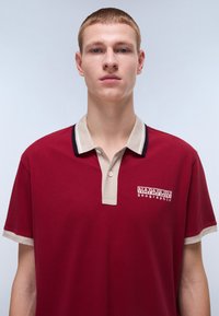 Polo shirt bordeaux con colletto e polsini beige a contrasto, con un logo ricamato in bianco sul lato sinistro del petto.