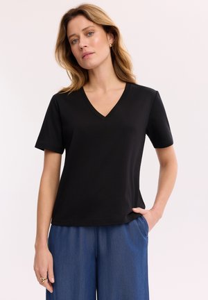 Kobieta z blond włosami do ramion, ubrana w czarną koszulkę z krótkim rękawem i dekoltem w serek oraz niebieskie spodnie, ręka w kieszeni na jednolitym tle.