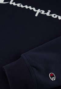 Námornícky sveter z mäkkej tkaniny s bielym logom "Champion", ribbovanými manžetami a malým emblémom s červenými, bielymi a čiernymi akcentmi.