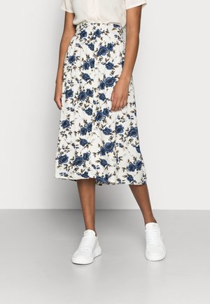 Kvinde iført en hvid midi-nederdel med blåt blomsterprint, en hvid top stoppet ned i nederdelen og hvide sneakers, stående foran en ensfarvet baggrund.