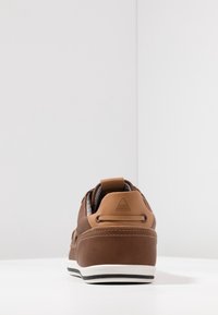Sneaker décontracté marron vu de dos avec languette de talon beige, détails cousus, et semelle blanche à rayures noires sur une surface blanche.