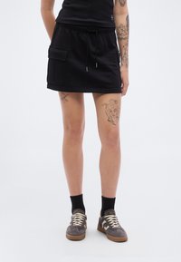 Μαύρη μίνι φούστα από βαμβάκι με ελαστική μέση, κορδόνι και πλαϊνές τσέπες cargo. Φορεμένη με γκρι αθλητικά παπούτσια και μαύρες κάλτσες.