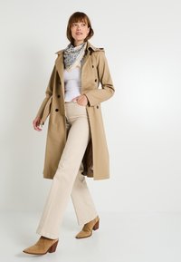 Gabardina beige con diseño de doble botonadura, botones negros y cintura ceñida con cinturón. Combinada con pantalones color crema y botas de tacón color beige.