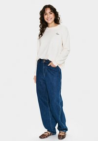Chemise crème à manches longues avec un petit logo rouge, associée à un jean en denim taille haute et des ballerines à motif léopard, affichant une coupe décontractée.