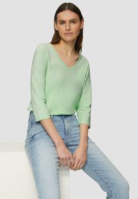 Lichtgroen V-hals lange mouwen top van zacht materiaal, gecombineerd met hoge taille lichtblauwe jeans, gepresenteerd tegen een neutrale achtergrond.