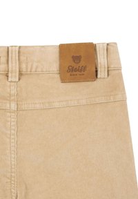 Beige korduroy kalhoty s koženou nášivkou s logem medvěda a nápisem "Steiff" na pase. Viditelné detaily šití.