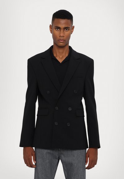 Blazer nero doppiopetto, design strutturato, ampi revers, tre bottoni, due tasche frontali, tessuto testurizzato, vestibilità leggermente oversize.