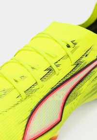 Kirkkaan keltainen jalkapallokenkä teksturoidulla päällisellä, mustilla yksityiskohdilla ja punaisilla korostuksilla. Varustettu perinteisillä nauhoilla ja virtaviivaisella muotoilulla.